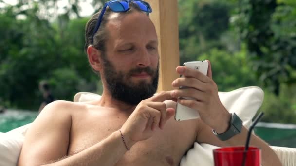 Homem usando smartphone enquanto deitado na cama de sol pela piscina — Vídeo de Stock