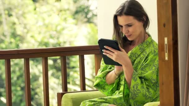 Vrouw in badjas met behulp van de kindle op terras — Stockvideo