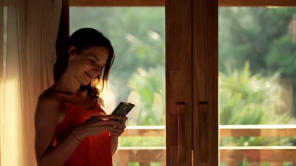 Mulher usando smartphone em pé pela janela — Vídeo de Stock