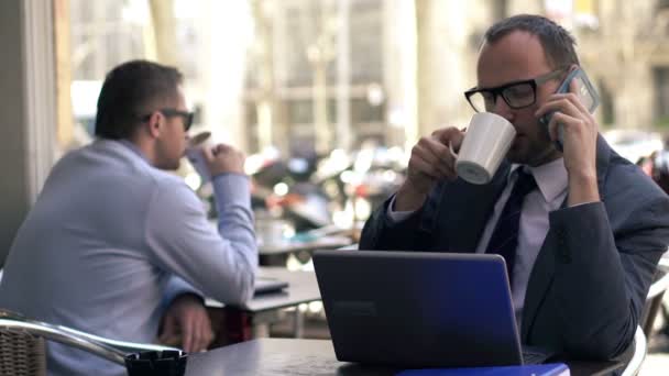 Empresario hablando por celular delante del portátil en la cafetería — Vídeos de Stock