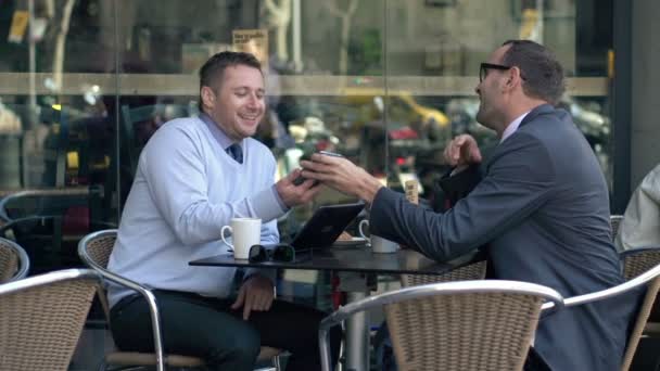 Два бизнесмена смотрят на что-то смешное на смартфоне в кафе — стоковое видео
