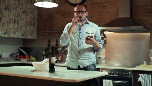 Молодой человек пишет смс на смартфоне и пьет красное вино на кухне — стоковое видео