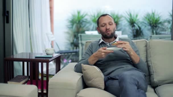 Hombre comiendo sándwich y tomando té mientras está sentado en el sofá en casa — Vídeos de Stock