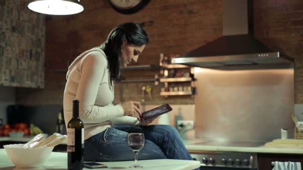 Молода жінка з планшетним комп'ютером п'є вино на кухні — стокове відео