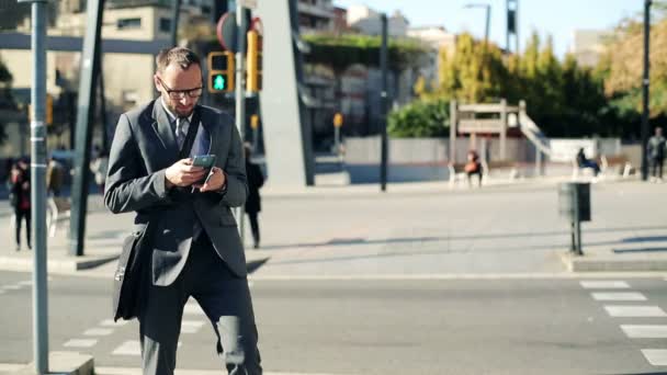 Молодой бизнесмен со смартфоном пересекает городскую улицу — стоковое видео