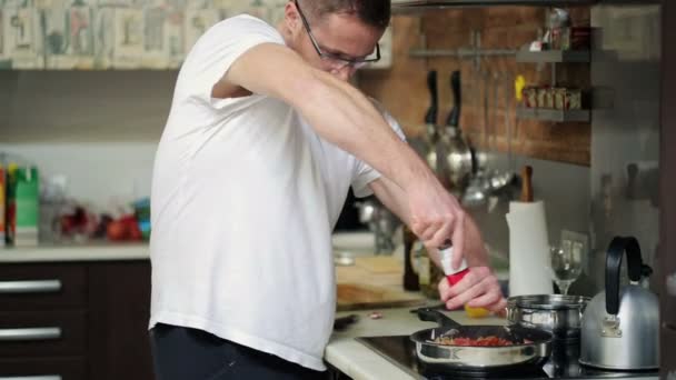 Jeune homme cuisine, ajouter des épices au repas de friture dans la cuisine — Video