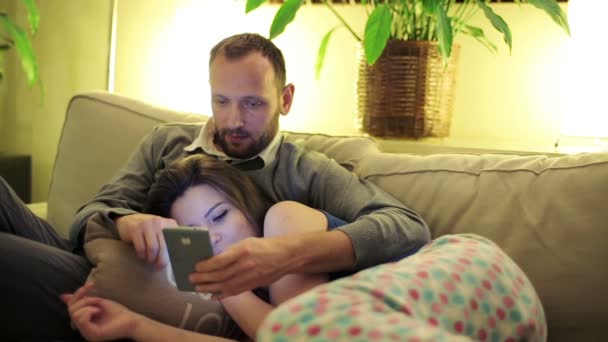 Νεαρό ζευγάρι με smartphone να καθίσει στον καναπέ στο σπίτι, νύχτα ώρα — Αρχείο Βίντεο