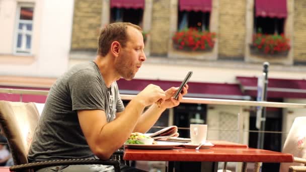 Мужчина пишет смс на смартфоне, ест сэндвич в ресторане — стоковое видео