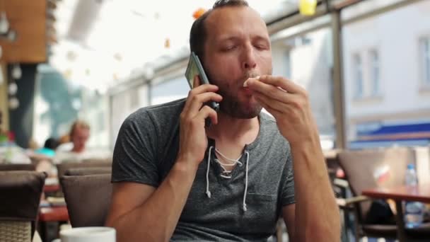 Νεαρός άνδρας που μιλάει στο κινητό, τρώει σάντουιτς στην καφετέρια — Αρχείο Βίντεο