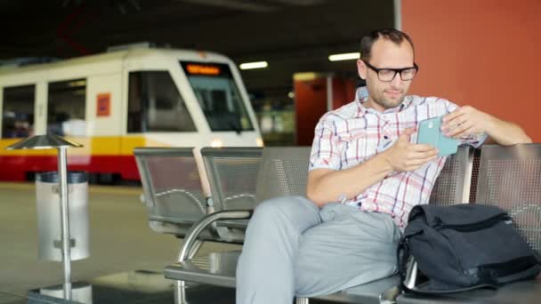 Młody człowiek z smartphone siedzi na dworcu — Wideo stockowe