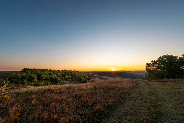 Mavi gökyüzünde turuncu bir parıltı, şafak söküyor. Sonbahar otları ve ormanlar güneş ışığını yansıtır.