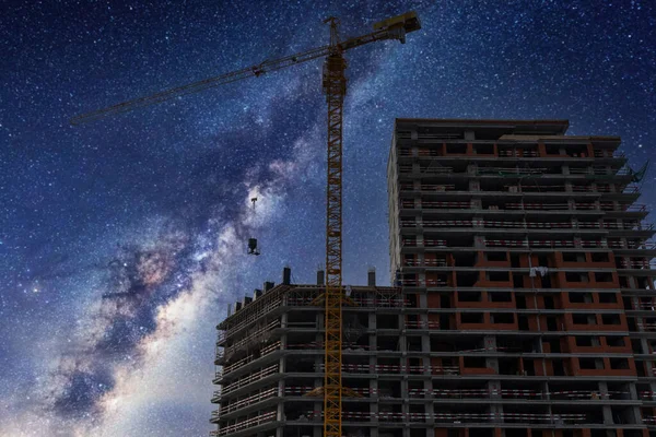 Tall tower crane and buildings and Milky way at night. A construction crane lifts a container for concrete with cement for unfinished house.