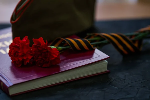 胜利日假期。黑黑的背景，圣乔治带和康乃馨躺在红皮书上. 图库照片