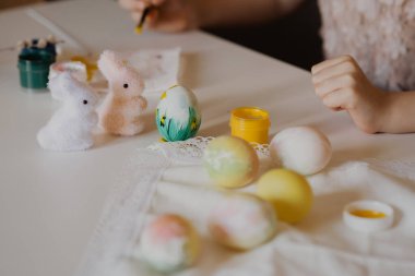 Paskalya çocuklarının yaratıcılığı. Küçük bir kız beyaz yumurta boyuyor. Diğer yumurta ve tavşanların yanında.