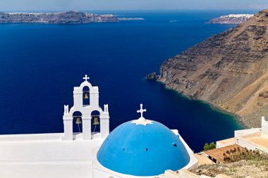 Santorini, Yunanistan: Mavi kubbeli resim gibi bir kilise ve çan kulesi, adanın ikonik kaldera manzarasını gösteren derin mavi Ege Denizi karşısında duruyor.