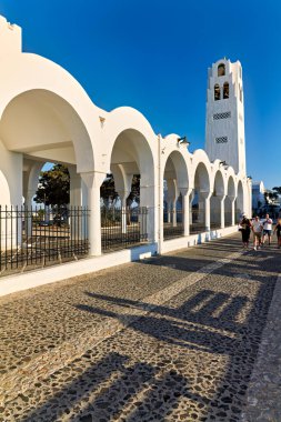Turistler, Yunanistan 'ın Santorini kentinde elinde çan kulesi ve kaldırım taşları olan klasik bir beyaz kilisenin önünden geçerek güneşli günün tadını çıkarıyorlar..
