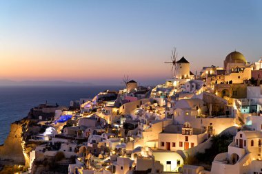 Oia, Santorini, Yunanistan alacakaranlıkta. Turistler, gün batımında Ege Denizi 'ne bakan aydınlanmış beyaz yıkanmış köy ve ikonik yel değirmenlerinin keyfini çıkarıyorlar..