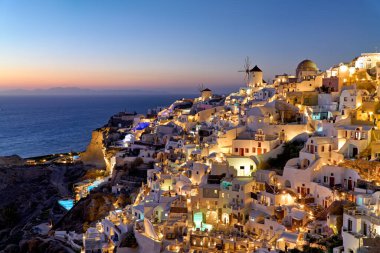 Yunanistan 'ın Santorini kentindeki Oia köyü alacakaranlıkta ışıklarla parlayarak, Ege Denizi' ne bakan ikonik beyaz binaları ve yel değirmenlerini gösteriyor..