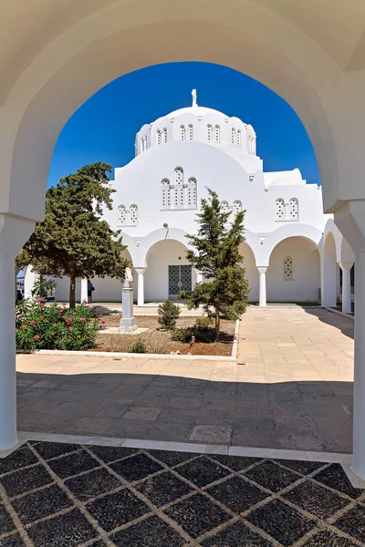 Kubbesi ve haçı olan el değmemiş beyaz bir kilise, tipik bir Cycladic mimarisi, güneşli bir avluda duruyor, kemer deliğinden görülüyor. Yunanistan.