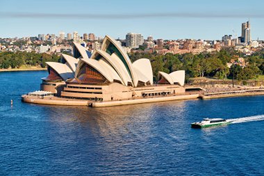 Sydney Opera Binası, Avustralya. Güneşli bir günde, ikonik bir simgenin önünde mavi sularda bir feribot yol alır..