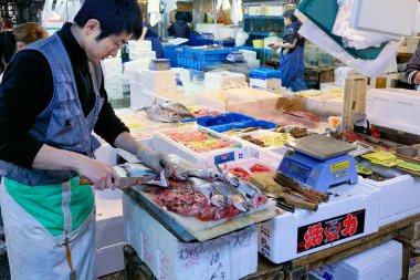 Bir satıcı Tokyo 'daki balık pazarında balık keser. Alıcılar çeşitli deniz ürünleri ve taze ürünlerle dolu tezgahlara göz atıyorlar..