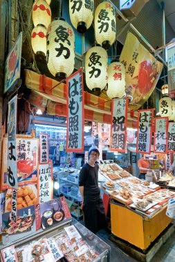 Müşteriler Kyoto 'daki Nishiki Pazarı' nı gün içinde keşfediyorlar. Tedarikçiler renkli tabelalar ve fenerler altında taze deniz ürünleri ve yerel atıştırmalıklar sergiliyorlar..