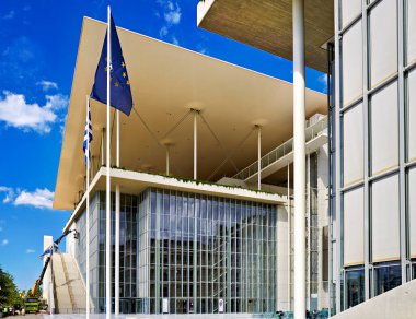 Yunanistan 'da Stavros Niarchos Vakfı Kültür Merkezi yer alıyor. Açık mavi bir gökyüzünün altında birçok bayrak duruyor. Ziyaretçiler binaya yaklaşıyor..