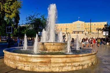 Ziyaretçiler, çeşmenin güneşli bir günde Yunan Parlamentosu binasının önüne su püskürttüğü Syntagma Meydanı 'nın keyfini çıkarıyorlar.