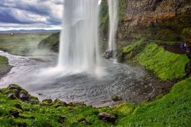 İzlanda 'da Seljalandsfoss' ta su çağlarken, turistler üssün yakınlarında yürüyüp fotoğraf çekiyorlar. Şelalenin etrafını yosun kaplıyor..