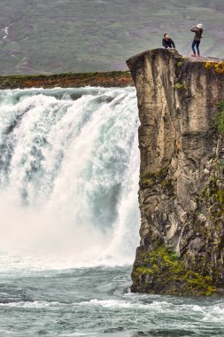 İzlanda 'da Godafoss şelalesinin yanında iki kişi kaya kenarında duruyor. Su güçlü bir şekilde akar ve sis yükselir..