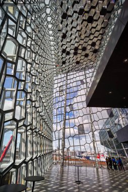 Ziyaretçiler Reykjavik 'teki Harpa konser salonuna parlak bir gökyüzünün altında bakıyorlar. Pencerelerin ve duvarların eşsiz tasarımı ilginç gölgeler yaratır..