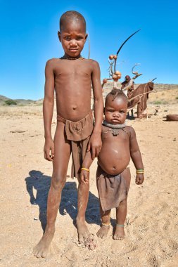İki Himba çocuğu Namibya 'nın Kunene bölgesinde kumlu zeminde duruyor. Geleneksel kıyafetler giyerler ve kültürlerini gösterirler..