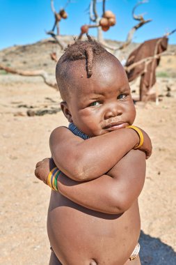Namibya 'nın Kunene bölgesinde kolları çaprazlanmış bir Himba çocuğu duruyor. Çevre kuru ve doğal. Arka planda birkaç kişi var..