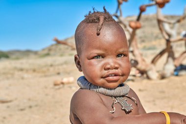 Himba 'lı bir çocuk Namibya' nın Kunene bölgesinde duruyor, gülümsüyor ve geleneksel mücevherleri bir çöl zeminine takıyor..