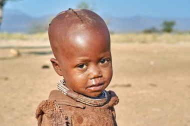 Namibya 'nın Kunene bölgesinde Himba köyünde bir çocuk duruyor. Çocuk basit bir ifadeyle doğrudan izleyiciye bakıyor..