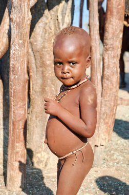 Himba köyünün dışında bir çocuk duruyor. Güneş köyde parlıyor. Çocuk merakla kameraya bakıyor..