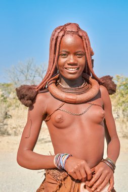 Himba 'lı bir kadın Namibya' nın Kunene bölgesinde güven içinde duruyor. Geleneksel takılar takıyor ve parlak güneş ışığında kameraya gülümsüyor..