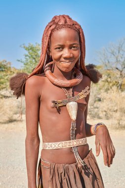 Namibya 'nın Kunene bölgesinde gülümseyen bir Himba kadını. Kuru bir manzarayla çevrili iken geleneksel giysiler ve mücevherler giyer..