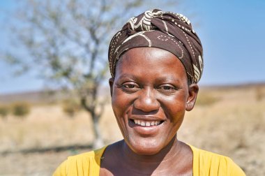Zemba Bantu etnik grubundan genç bir kadın, Kunene Bölgesi 'nde, temiz bir gökyüzünün altında, karalar ve ağaçlarla çevrili bir şekilde gülümsüyor..