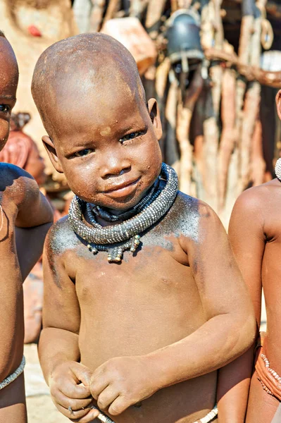 Namibya 'nın Kunene bölgesinde Himba köyünde bir çocuk duruyor. Çocuk geleneksel takılar takıyor ve yüzünde neşeli bir ifade var..
