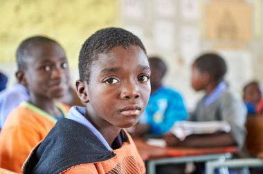 Bir erkek öğrenci okul zamanı, Kavango Bölgesi, Rundu 'da bir sınıfın masasında oturuyor. Arka planda diğer öğrenciler var.