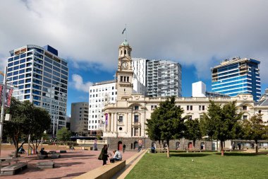 Aotea Meydanı, Auckland 'daki belediye binasına ev sahipliği yapıyor. Modern binalar arka planda bulutlu bir gökyüzünün altında yükselirken insanlar çimenlerde dinleniyor..