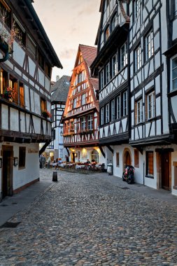 İnsanlar Petit France, Strasbourg 'da kereste çerçeveli evlerin yanındaki masalarda oturuyorlar. Güneş arka planda batıyor..