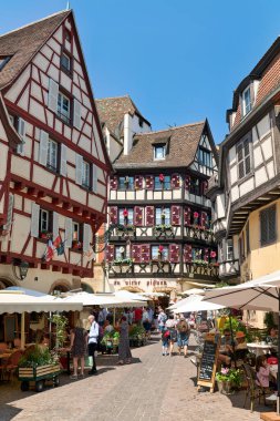 İnsanlar eski Colmar kasabasında yürüyor, kereste çerçeveli evleri görüyor ve parlak şemsiyeler altında market tezgahlarını keşfediyorlar..