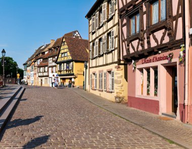 Ziyaretçiler Petite Venise 'deki kaldırım taşı caddesinde yürürken güneşli bir günde Alsace, Colmar' ın kereste çerçeveli evlerinin keyfini çıkarıyorlar..