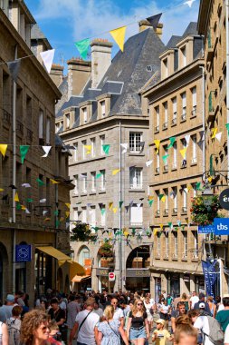 İnsanlar Saint Malo 'daki eski kasabanın sokaklarında yürüyor. Mağazalar ve kafeler ziyaretçileri karşılarken renkli bayraklar yukarıda asılı.