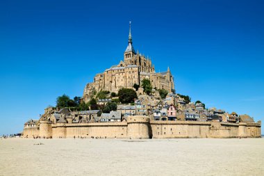 Mont Saint Michel dalgalar çekildikçe ayakta duruyor. Ziyaretçiler kumlu kıyıları keşfeder. Mavi gökyüzü şehir simgesi için açık bir zemin oluşturuyor.