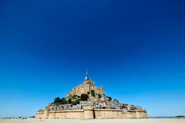 Mont Saint Michel alçak gelgit sırasında açık mavi bir gökyüzüne karşı dimdik ayakta duruyor. Ziyaretçiler kumlu sahil boyunca yürüyor, bölgeyi keşfediyorlar..
