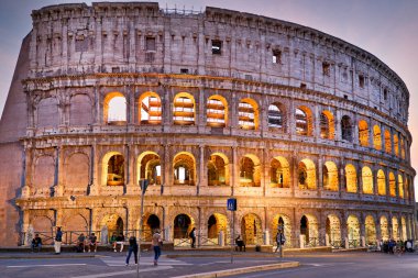 Roma 'daki Kolezyum' un yakınında toplanan insanlar akşam saatlerinde aydınlanıyor. Tarihi yapı, büyük kemer ve taş duvarlarını gösteriyor..