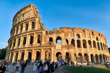 İnsanlar Roma 'daki Kolezyum' un etrafında yürüyorlar, açık mavi gökyüzünün altında, ünlü antik bir yapı. Burası popüler bir turistik yerdir..